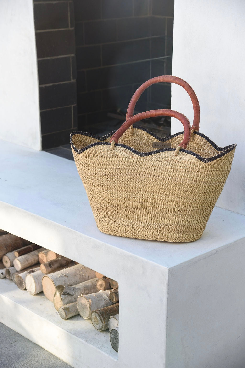 Woven basket with leather handle on a white outdoor fireplace next to stacked logs.