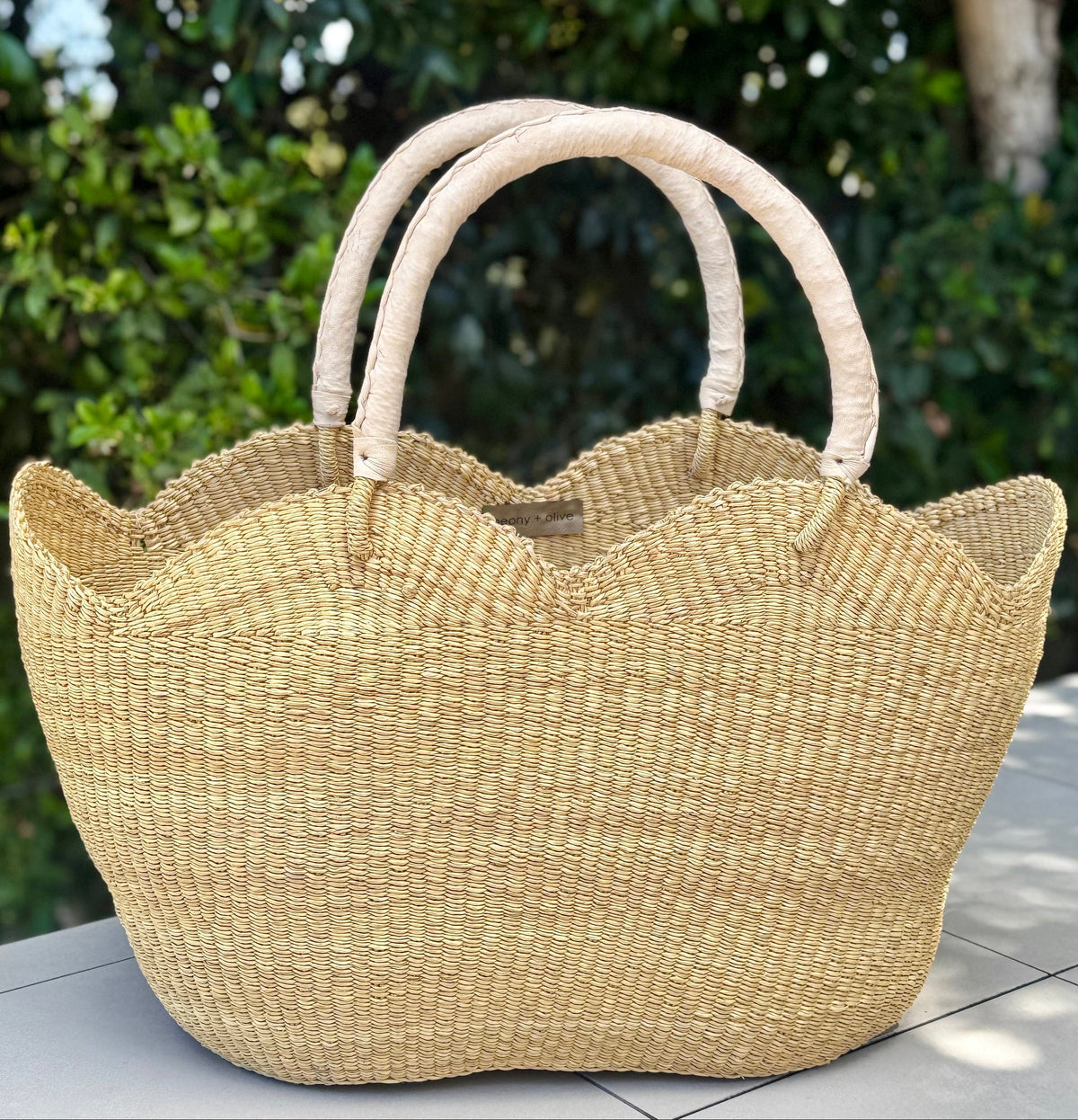 A natural woven straw tote bag with a scalloped edge and two loop handles, resting on a tiled patio with a blurred green garden background.