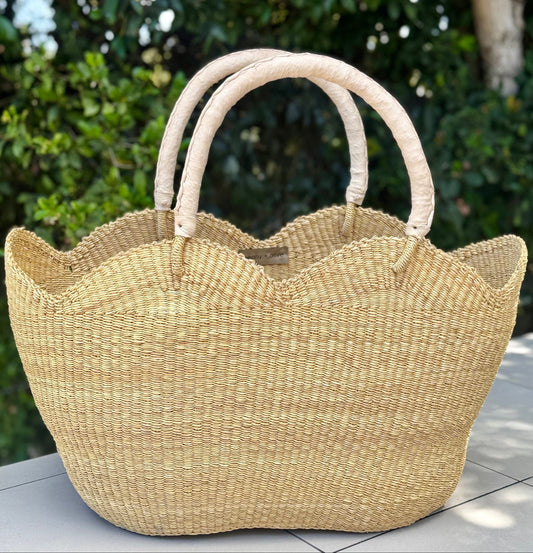 A natural woven straw tote bag with a scalloped edge and two loop handles, resting on a tiled patio with a blurred green garden background.