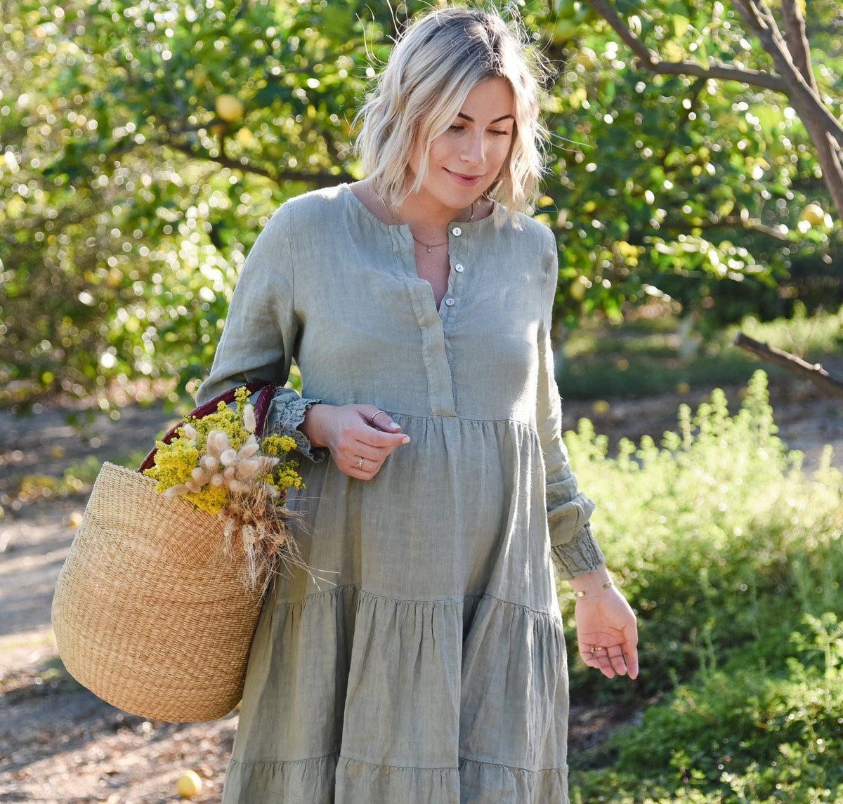 A woman stands outdoors in a sunlit orchard wearing a long-sleeve, tiered linen maxi dress in a muted sage green. She is holding a large woven U shopper basket filled with yellow flowers.