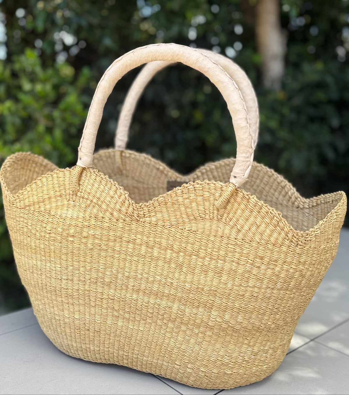 A natural woven straw tote bag with scalloped edges and two top handles, placed on a tiled surface with a blurred green garden background.