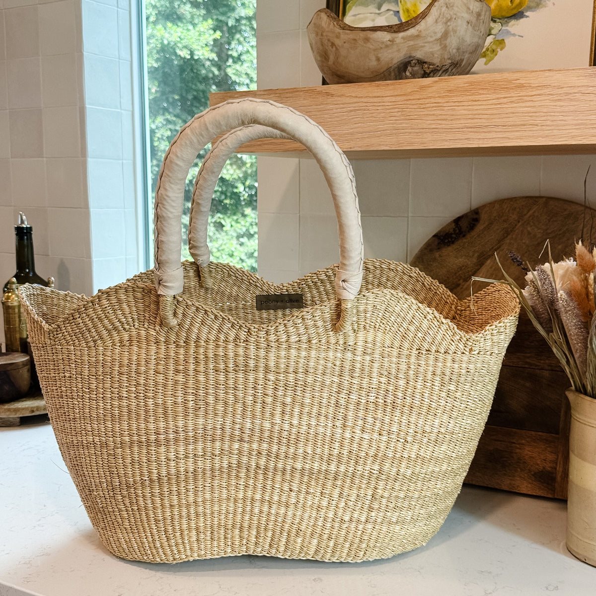 A natural woven tote bag with two curved  leather cream handles sits on a white kitchen countertop. The background shows a tiled wall, a wooden shelf with decorative items, and a window looking out to green foliage.