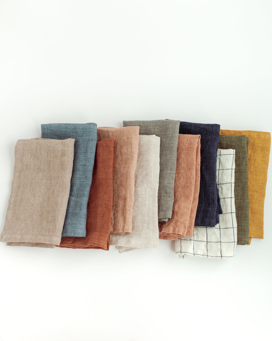 A collection of stone washed linen hemmed dinner napkins in various colors, neatly folded and displayed on a white background.